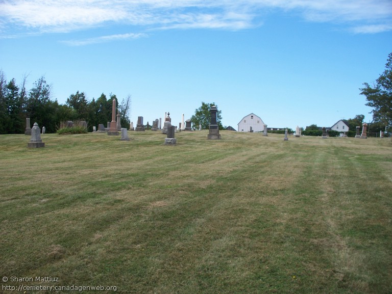 Wide view of Konkle-Marlatt Cemetery: scattered old headstones on a mown hillside with a white barn beyond