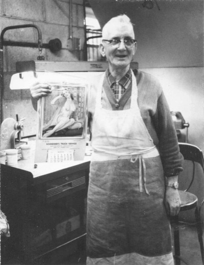 George D. Malott, older, in glasses and a work apron in a machine shop, smiling and holding up a pin-up girl calendar from Schneider&rsquo;s Truck Service