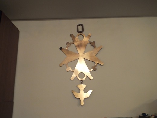 A large brass Huguenot cross hanging on a pale wall: a Maltese cross with heart-shaped openings and a dove suspended below