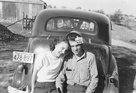 Wanda and Leroy Malott, smiling and leaning together against the trunk of a 1930s car with a 1951 Kentucky license plate, a wooden barn behind them