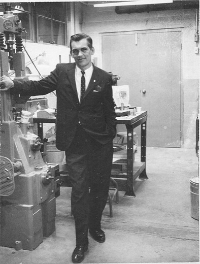 Leroy Joseph Malott in a dark suit and tie, leaning on a drill press in a machine shop, a workbench and steel door behind him