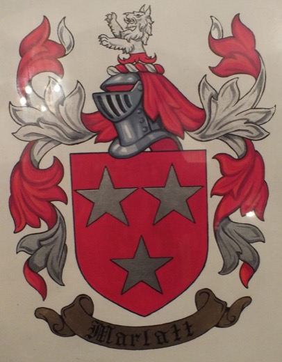 A hand-painted coat of arms: a red shield with three silver stars under a steel helmet and a rearing silver beast, red and silver mantling, and a scroll reading Marlatt