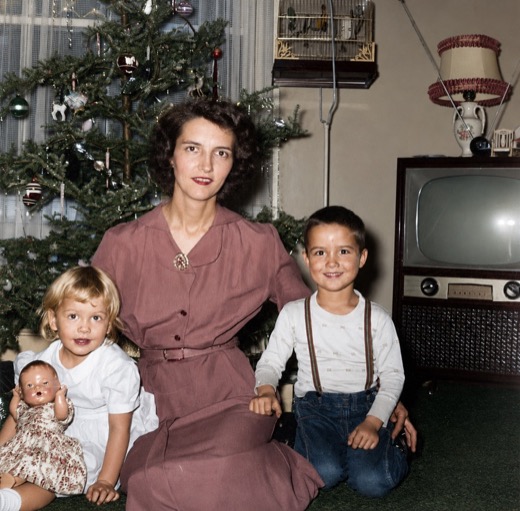 Wanda Malott in a rose-colored dress, kneeling in front of a Christmas tree with a young blond girl holding a doll and a smiling boy in suspenders, a television set beside them