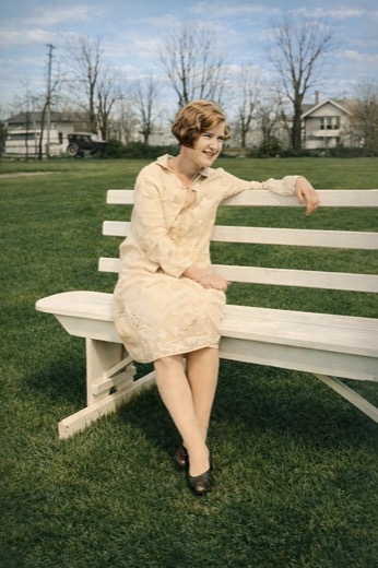 Mildred Adams as a young woman with bobbed hair and a cream lace dress, smiling on a white park bench, with houses and an early automobile behind her