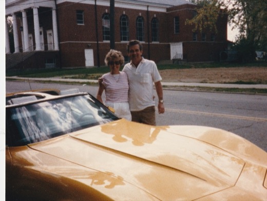 Robert E. Malott and his wife standing beside a gold Corvette Stingray convertible on a street near Norwood High School, a brick building with white columns behind them