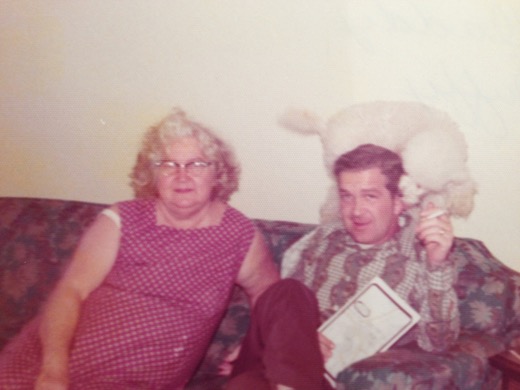 Mildred Malott in glasses and a pink print dress seated on a floral couch beside her son Robert, who holds a cigarette and a book, with a white poodle perched on the couch back behind him