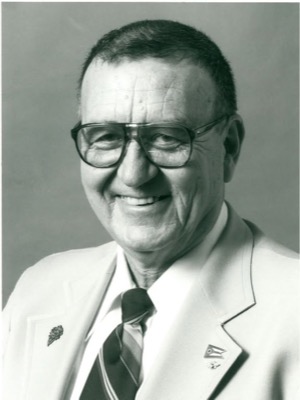 Black-and-white portrait of a smiling older man with short hair and large glasses, in a light jacket and striped tie, with an Ohio flag pin and another small pin on his lapels