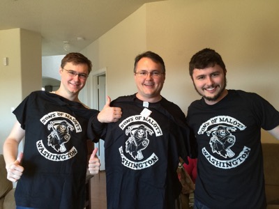 Three smiling men holding up black T-shirts printed Sons of Malott, Washington, with a hooded skeleton holding a scythe